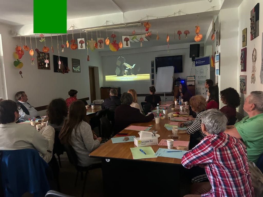 Participanți la o masă lungă ascultă o facilitatoare care prezintă slide-uri proiectate, lângă bannerul RE-CONECTAT