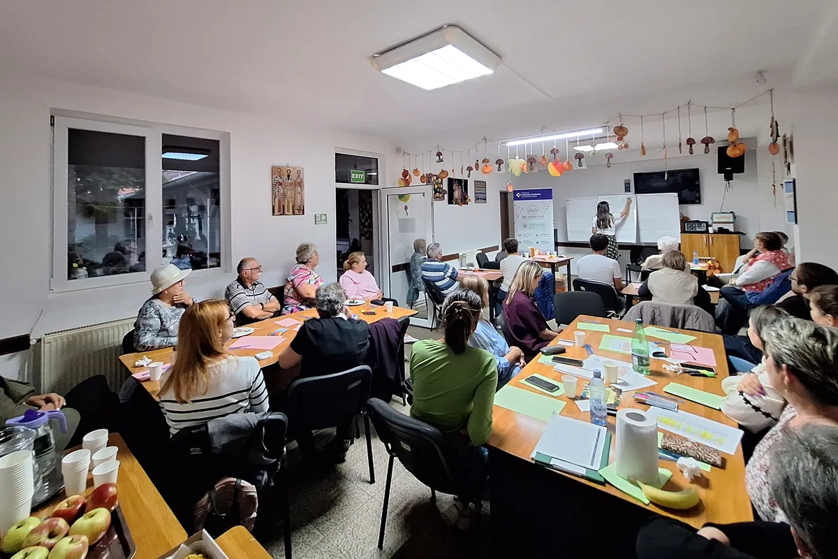 Adulți așezați la mese într-o sală decorată cu lucrări de toamnă urmăresc o facilitatoare care scrie pe flipchart
