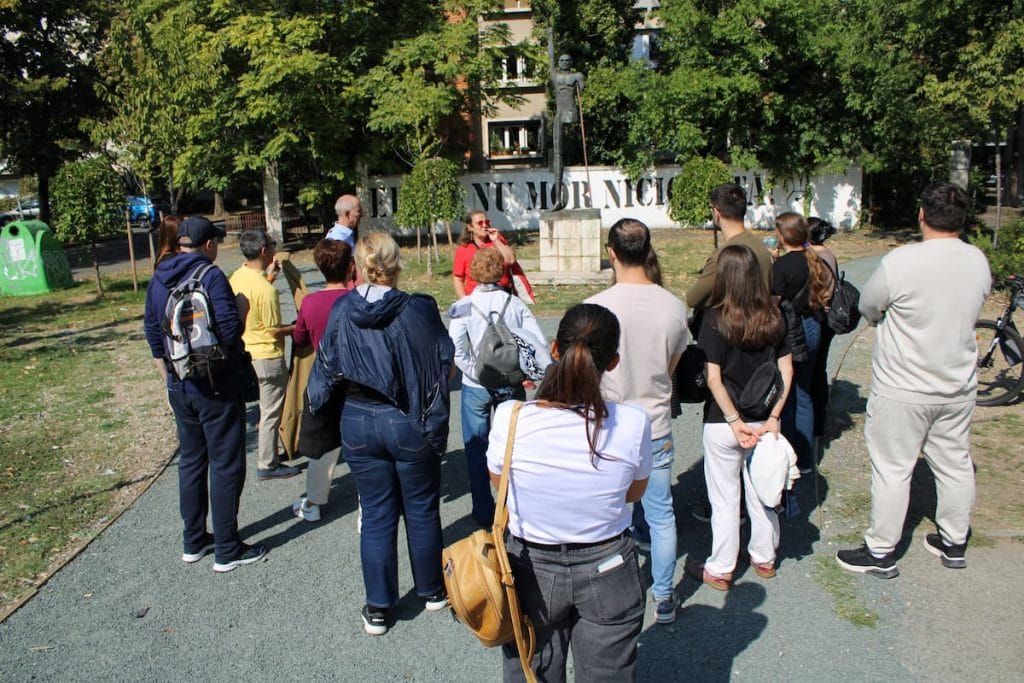 Grup de participanți adunat în semicerc pe o alee cu copaci, ascultând explicațiile unei ghide